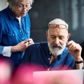 retired gentleman on computer with nervous lady looking over shoulder