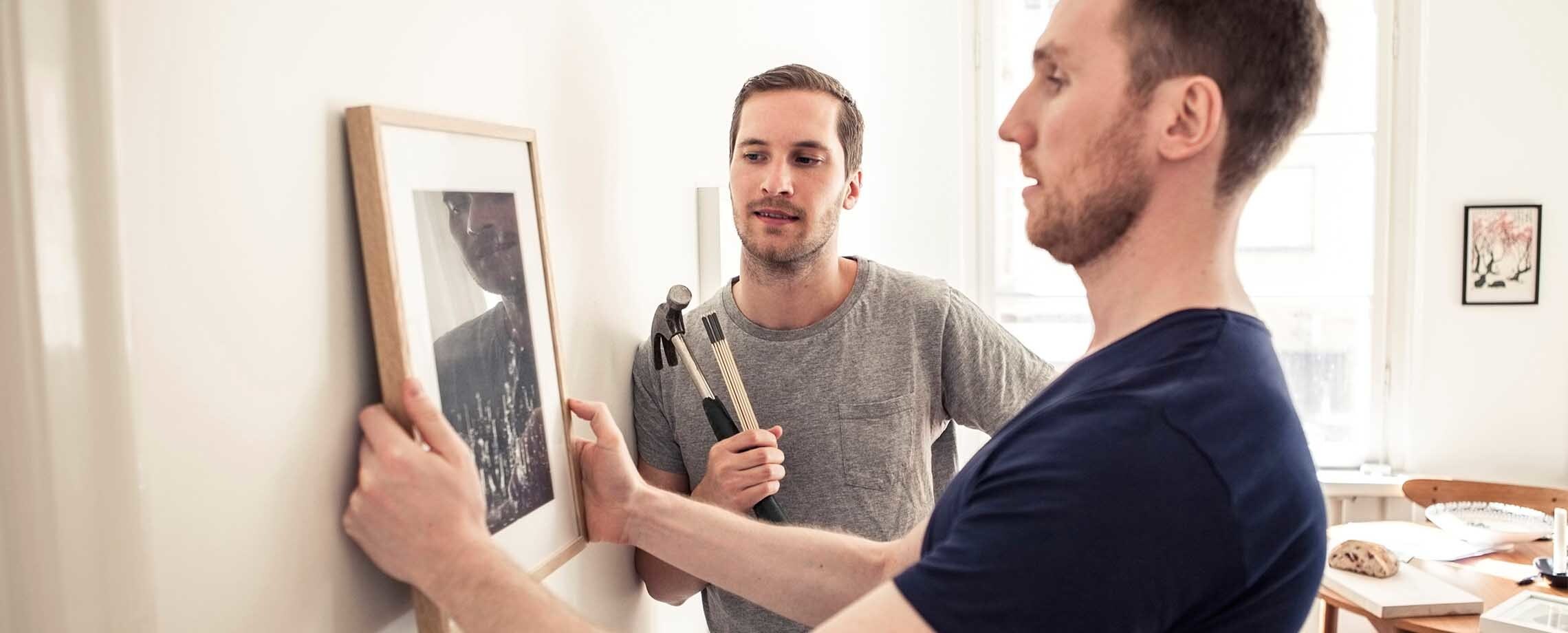 Two young men with hammer and ruler hanging photo on wall