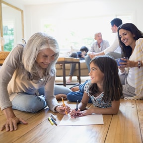 Grandmother, with granddaughter coloring on the floor with family in the background