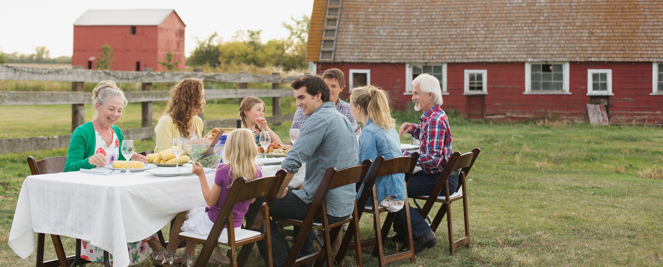 Seven family members of different generations having dinner outdoors in front of farmhouse