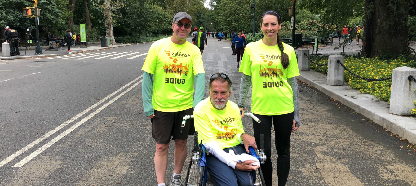 A man and woman standing being a man in a wheelchair, all wearing green/yellow “Guide” shirts by the side of a road in a park