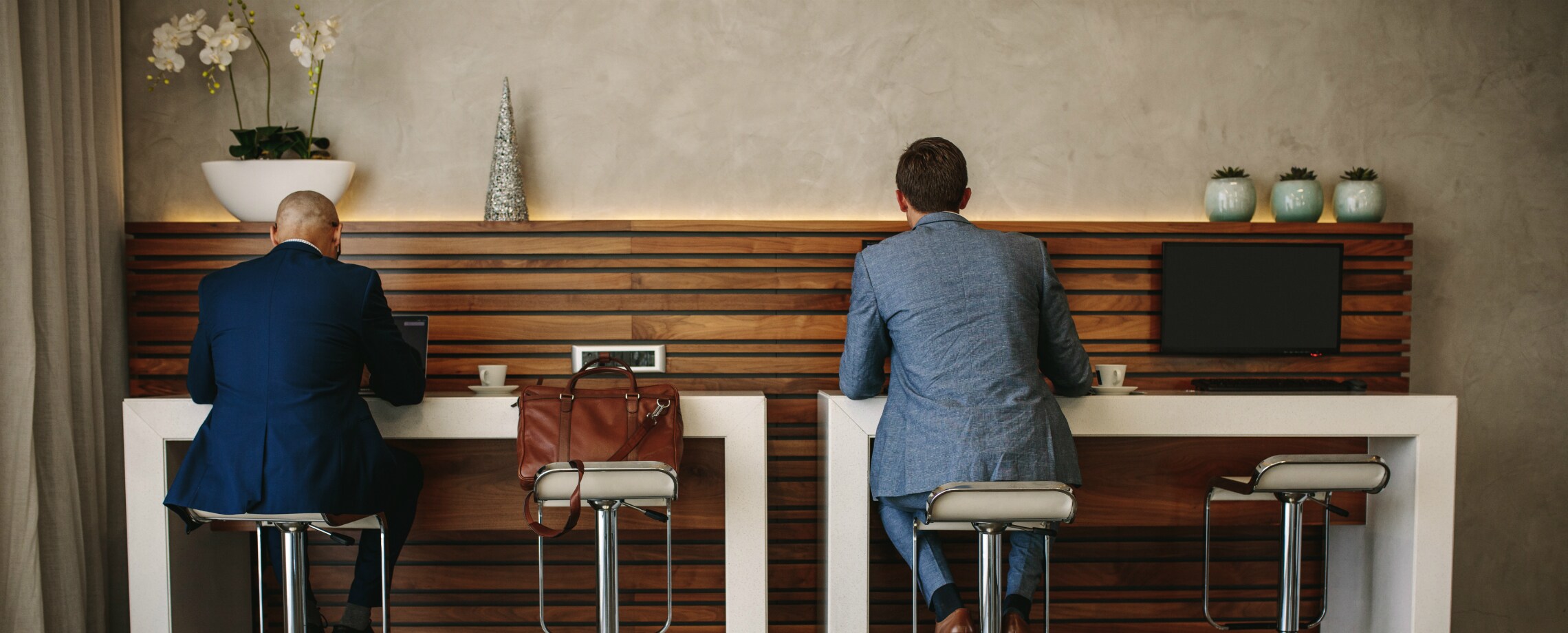Two men in suits using computers against a wall with a bag on stool between them