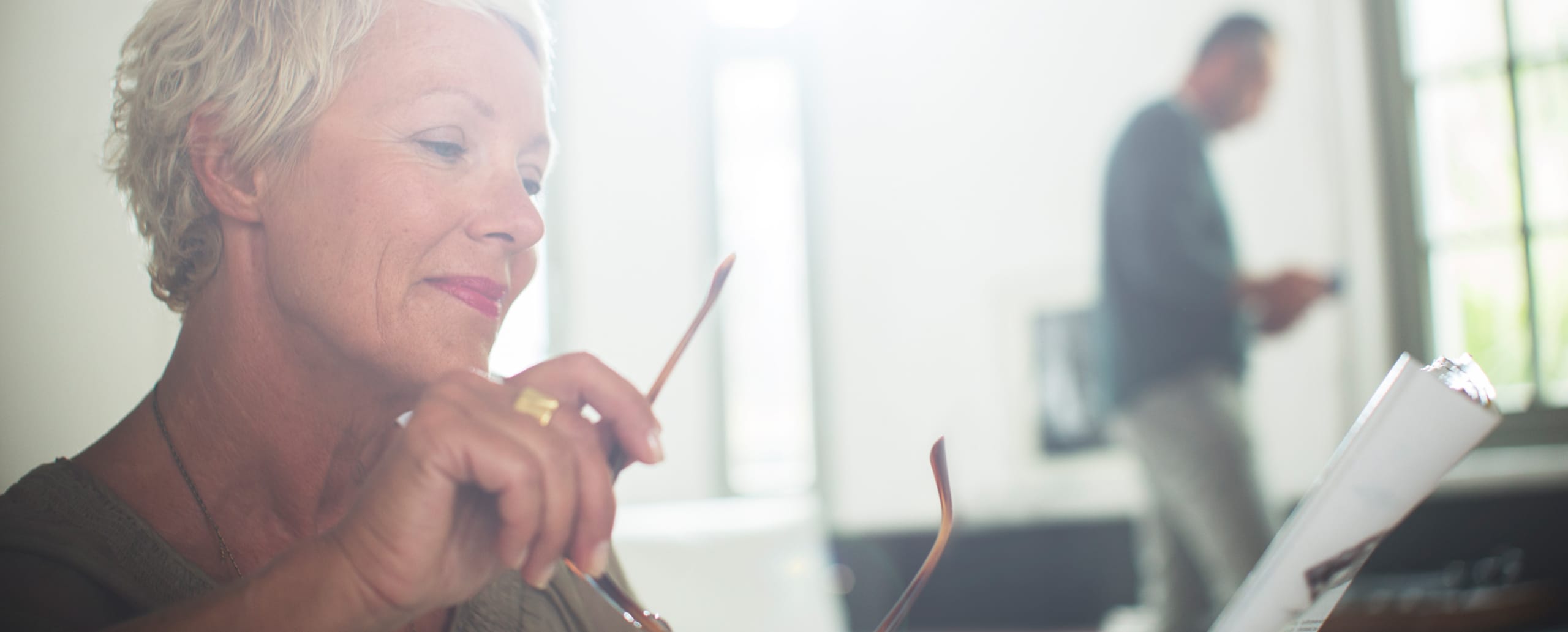 Caucasian woman holding glasses reading a magazine with man in the background