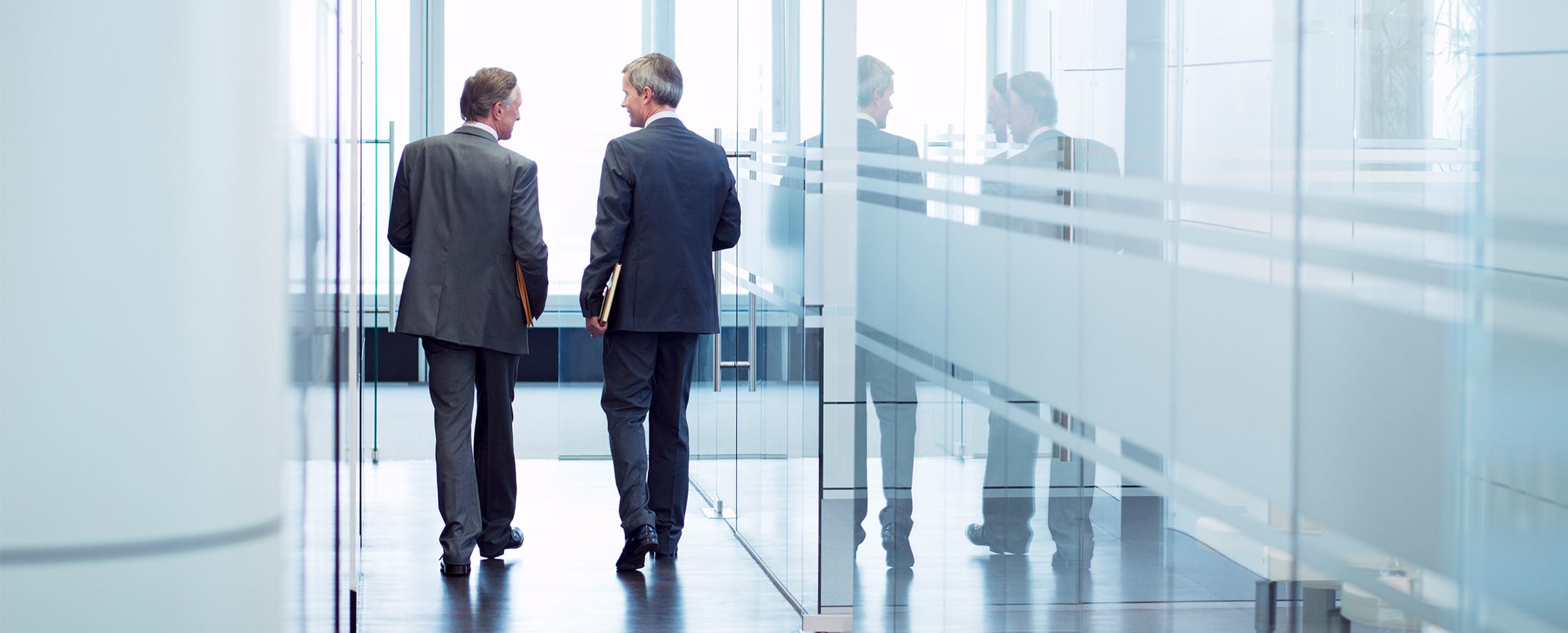 Two businessmen walking down glass-walled hall in office