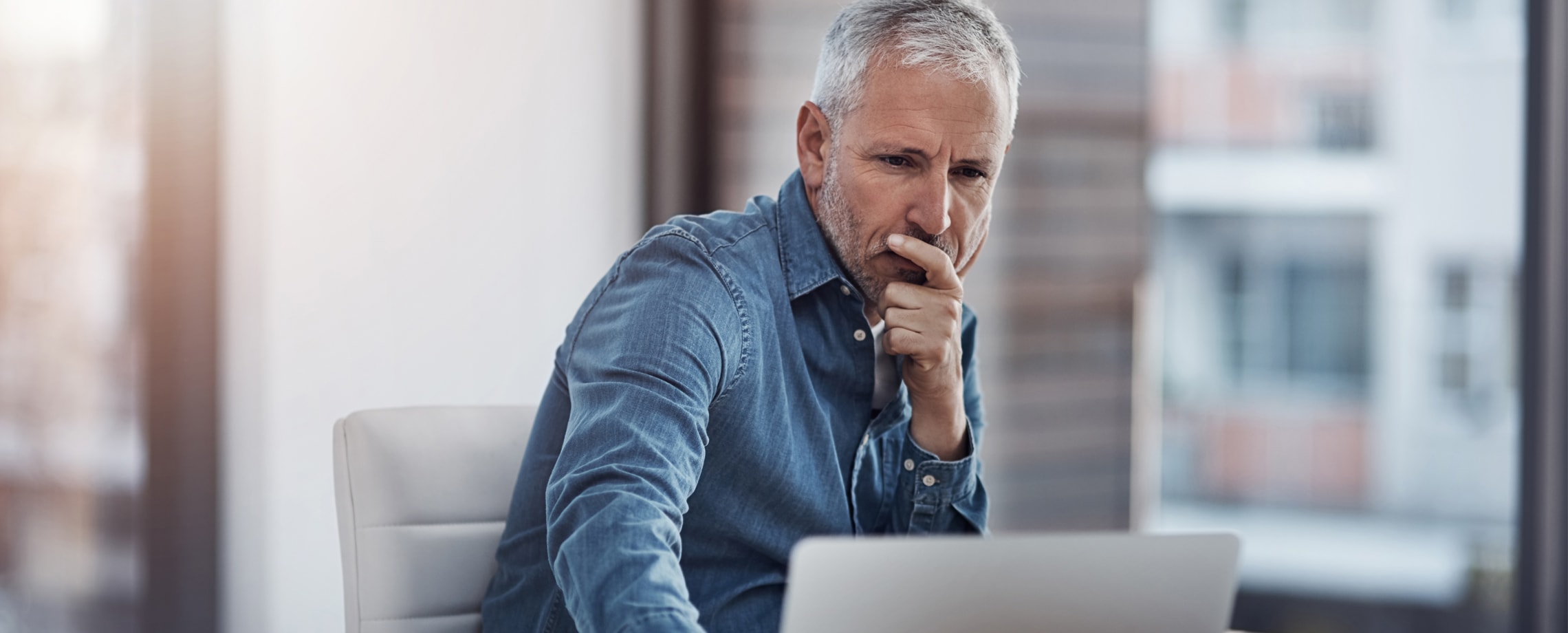 White haired bearded Caucasian male in denim shirt looking at laptop