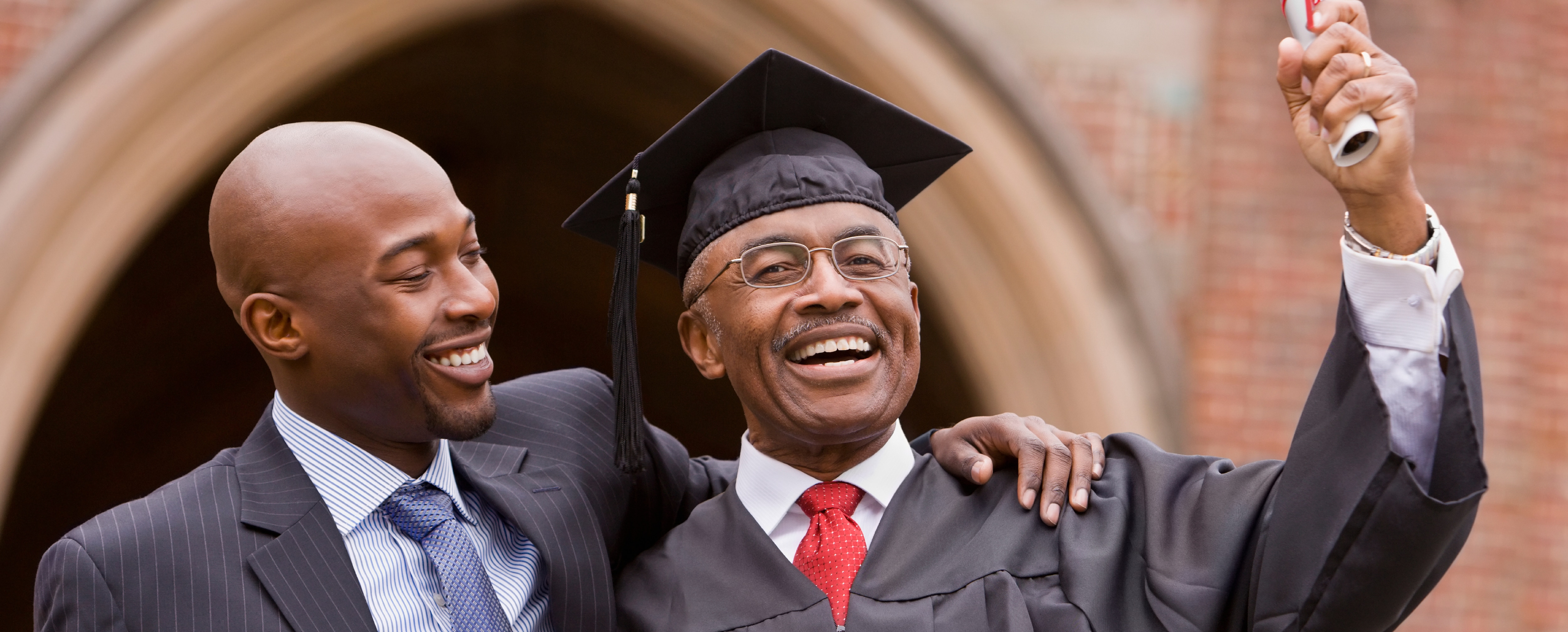 Older African-American man in cap and gown holding diploma with smiling son's arm around his shoulders