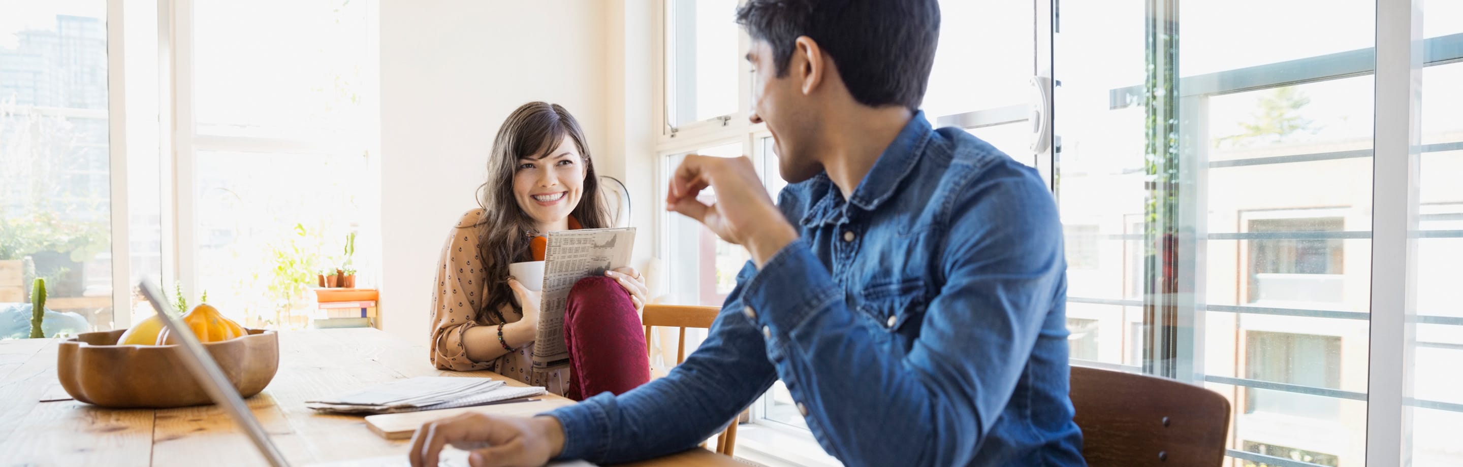 Young couple sitting at kitchen table, the woman is holding a newspaper smiling looking at the man who is on a laptop