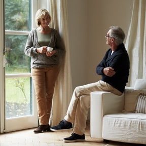 Retirement aged Caucasian couple with woman holding a coffee cup standing smiling down at husband sitting on arm of couch