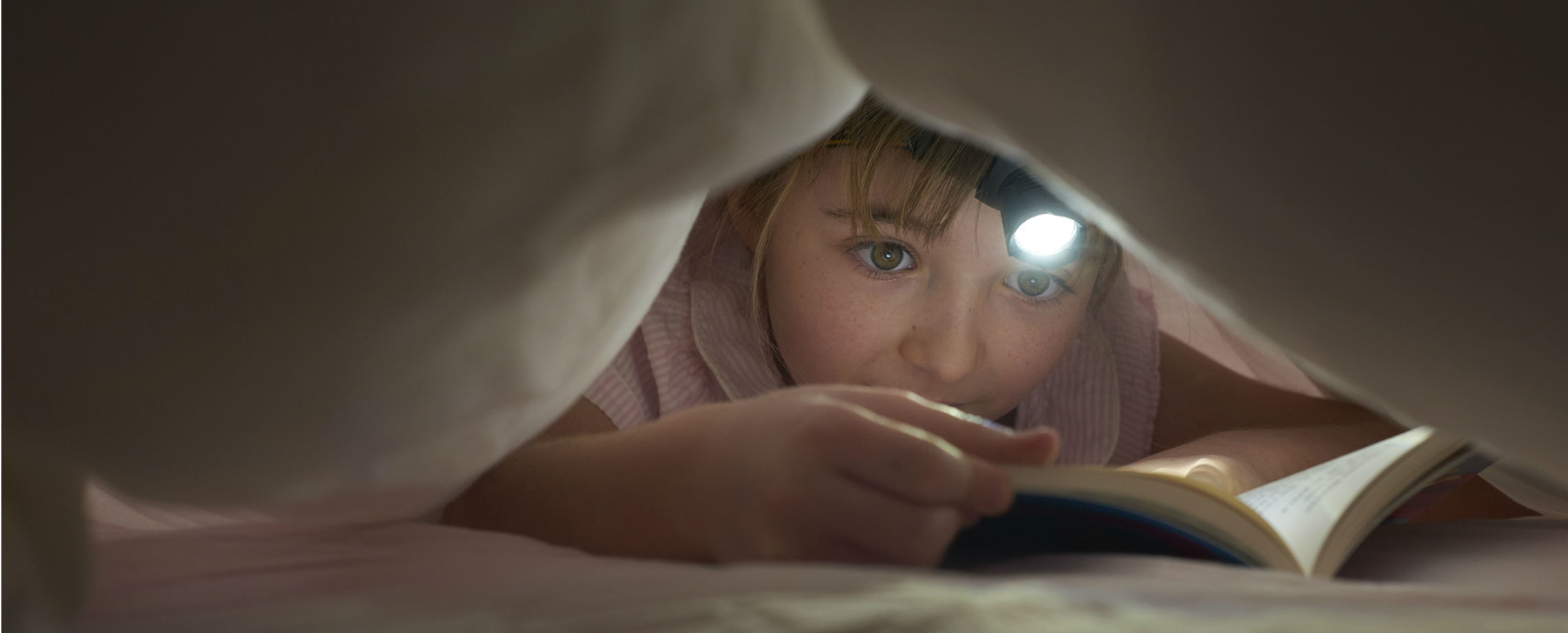 Caucasian child with a miner's headlamp reading under the covers