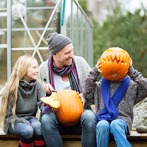 Man in sweater carving pumpkins with two children outside; one child is holding carved pumpkin in front of their face