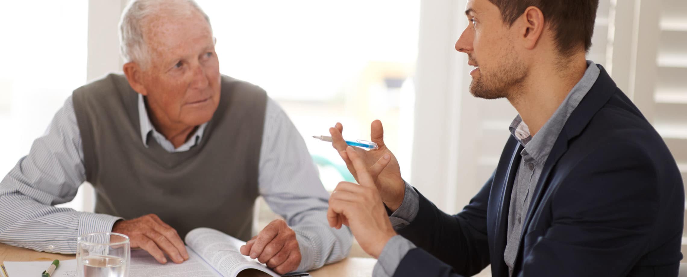 Retirement aged Caucasian man sitting at table with an open book listening to a younger man in sportscoat explaining something