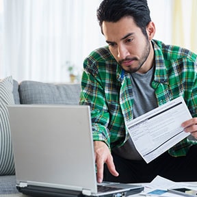 Bearded man at home, holding paperwork and looking at laptop