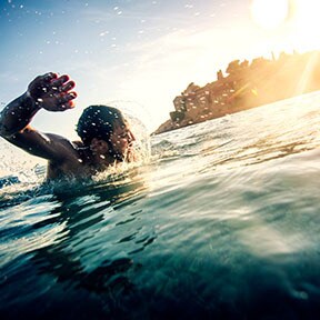 Man swimming in ocean with sun shining and buildings on the coast
