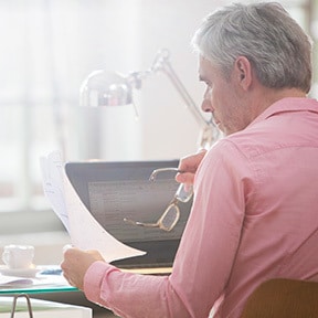 Man at computer holding and reading a paper sheet with glasses in his hand