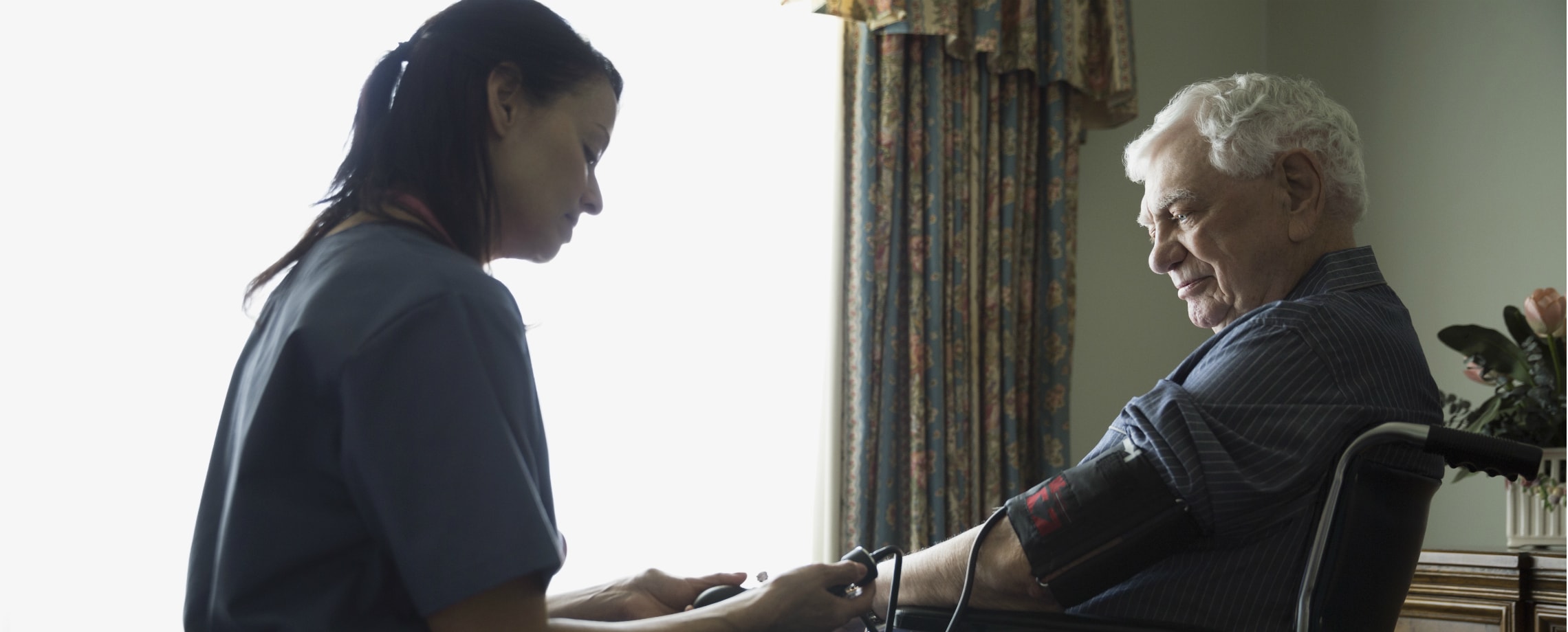 Nurse taking blood pressure of older grey haired man sitting in wheelchair