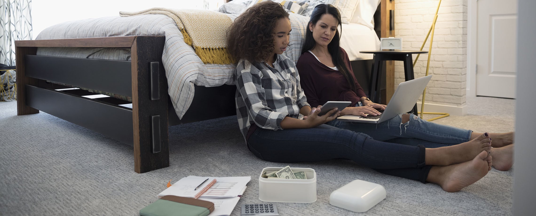 Mixed teenaged girl and Caucasian mother sitting on floor of bedroom with laptop and box with dollar bills in it