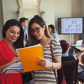 Many students in a room with two young women discussing papers they are holding