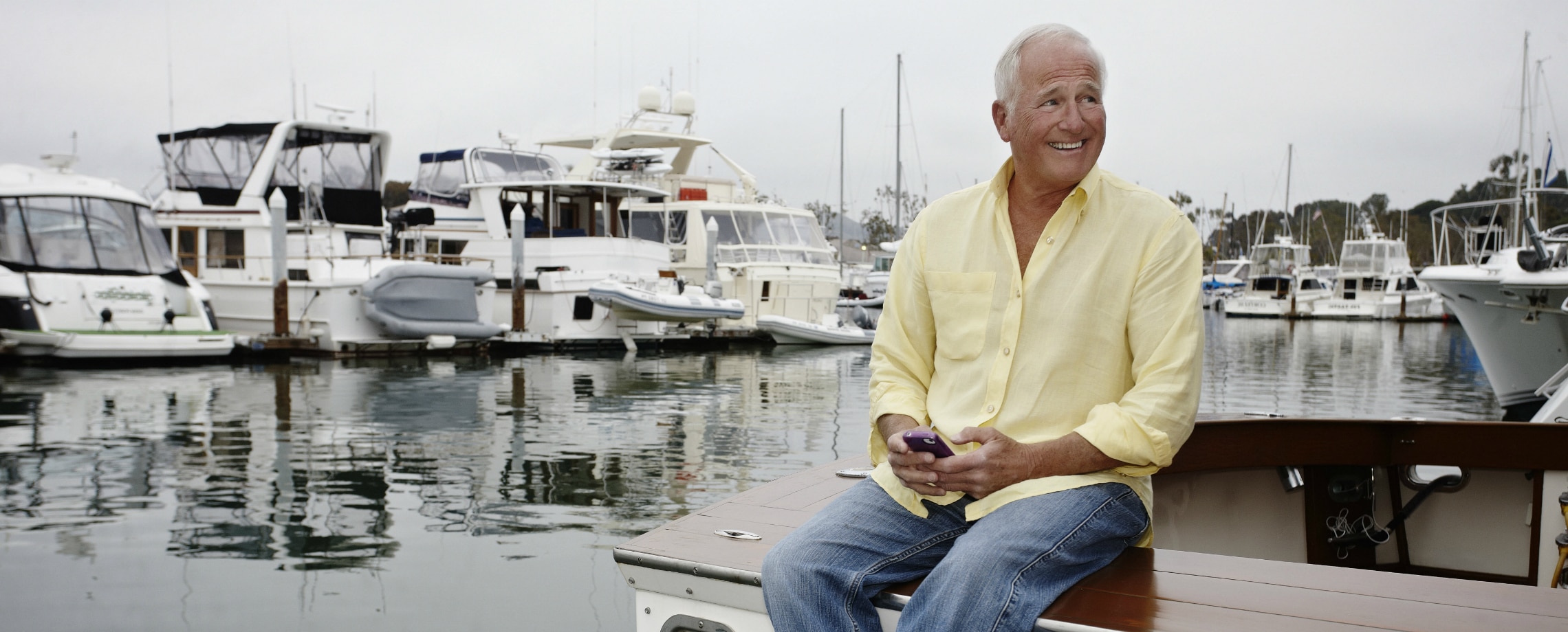 Older Caucasian man sitting on a boat with no shoes and his pants rolled up holding a cellphone