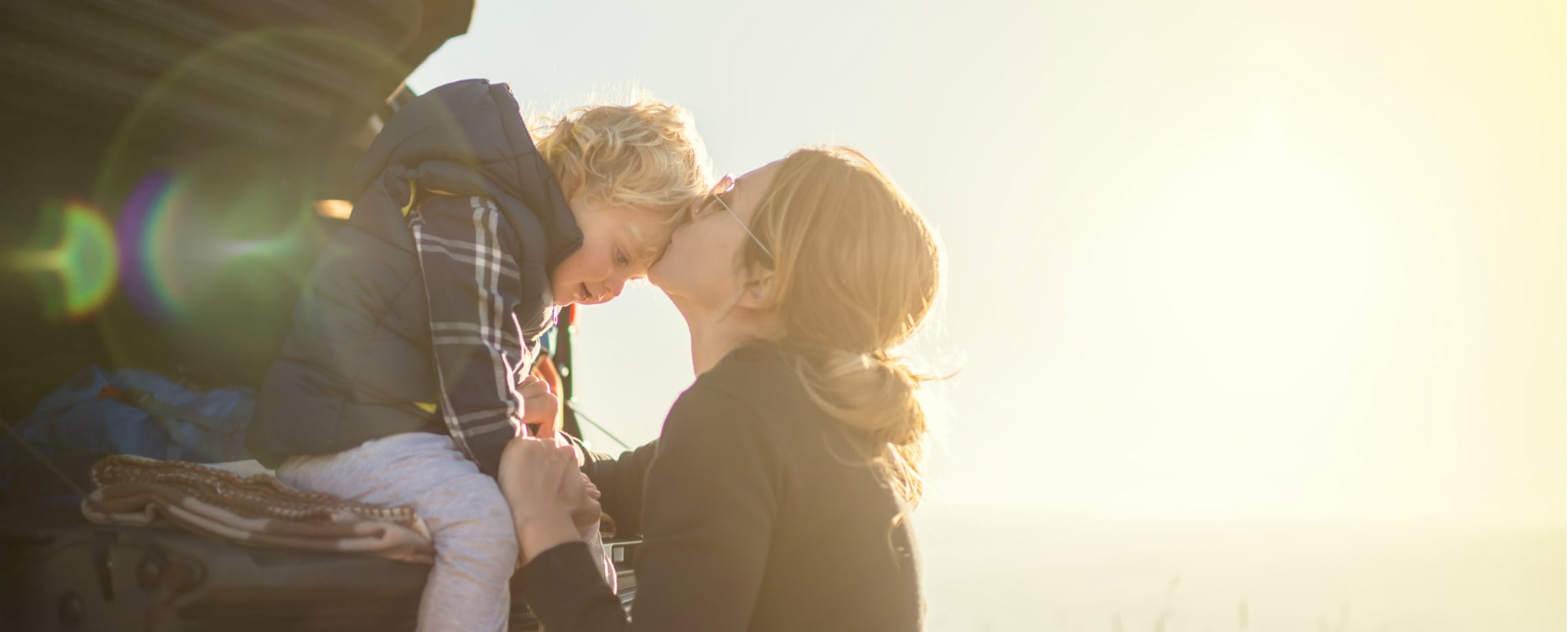 Close up of woman kissing her toddler son's forehead as he sits on tailgate of vehicle