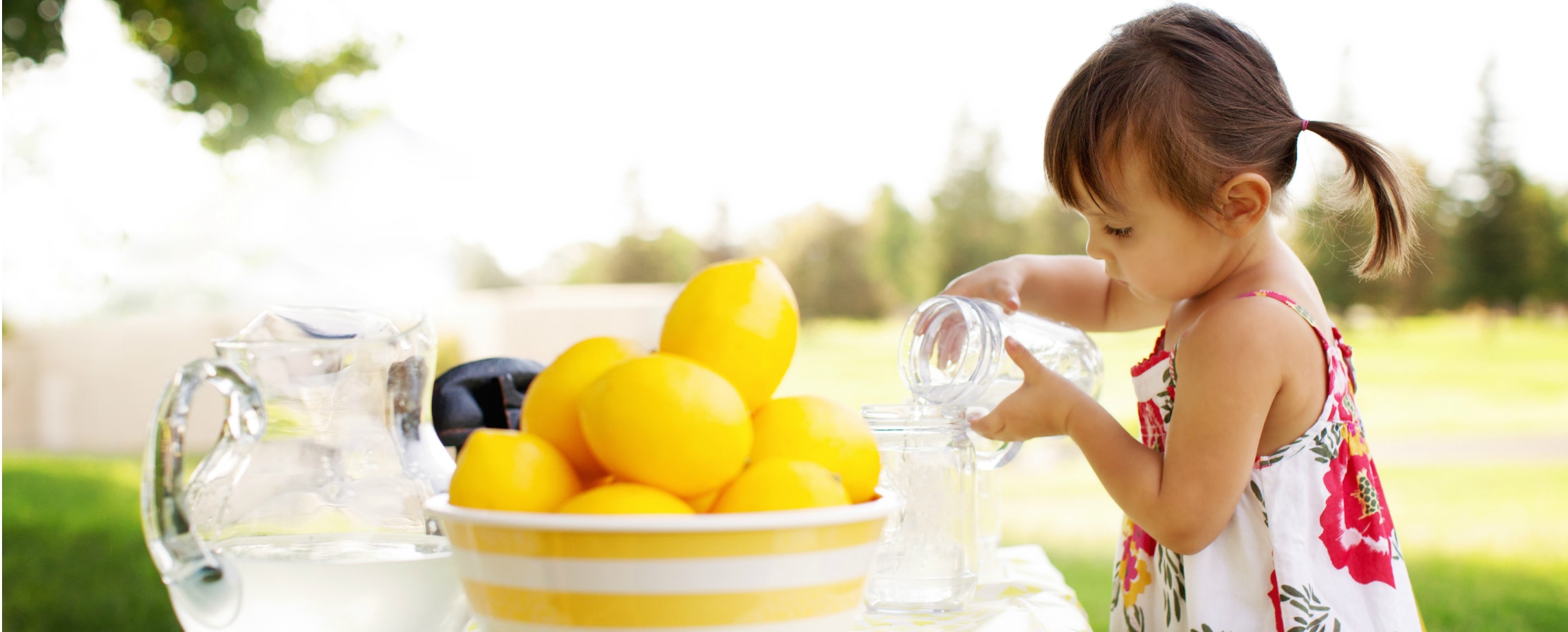 Very young girl pouring water from one mason jar to another with bowl of lemons on the table