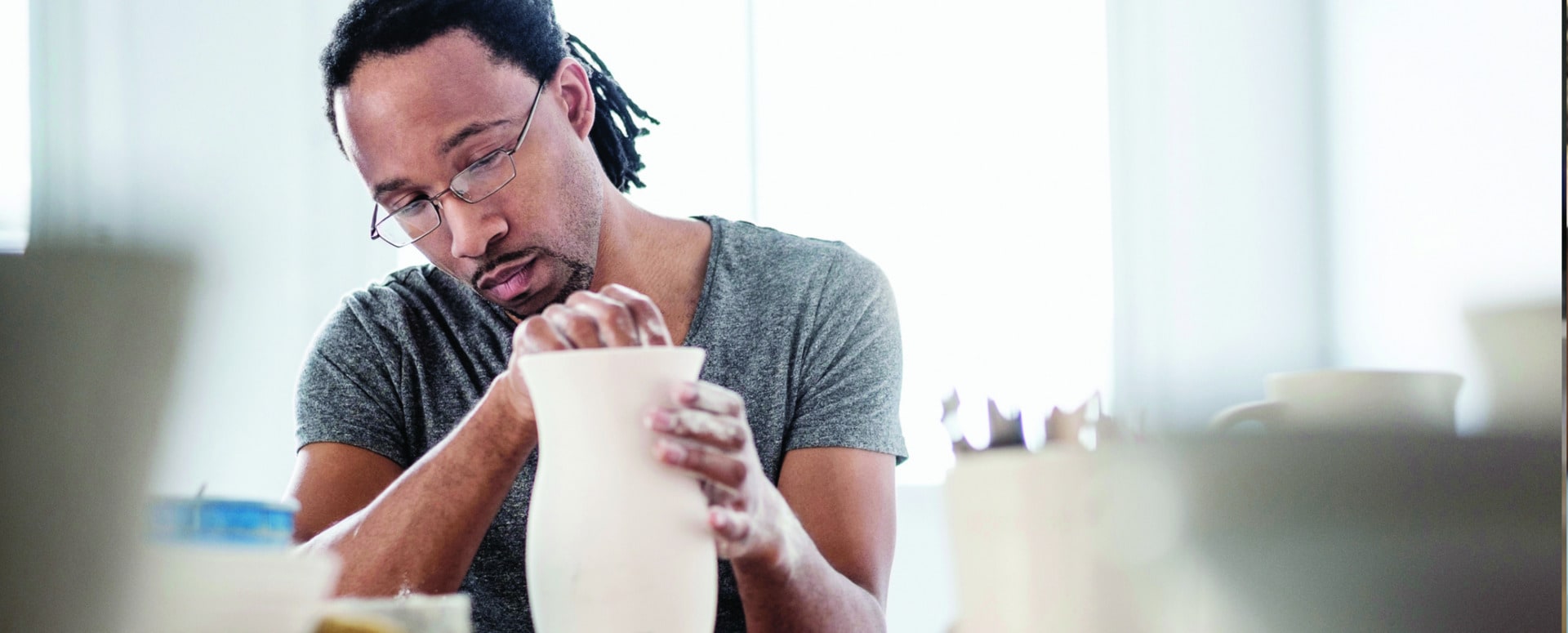 African-American male with dreads, a gotee and glasses sculpting a vase on a pottery wheel