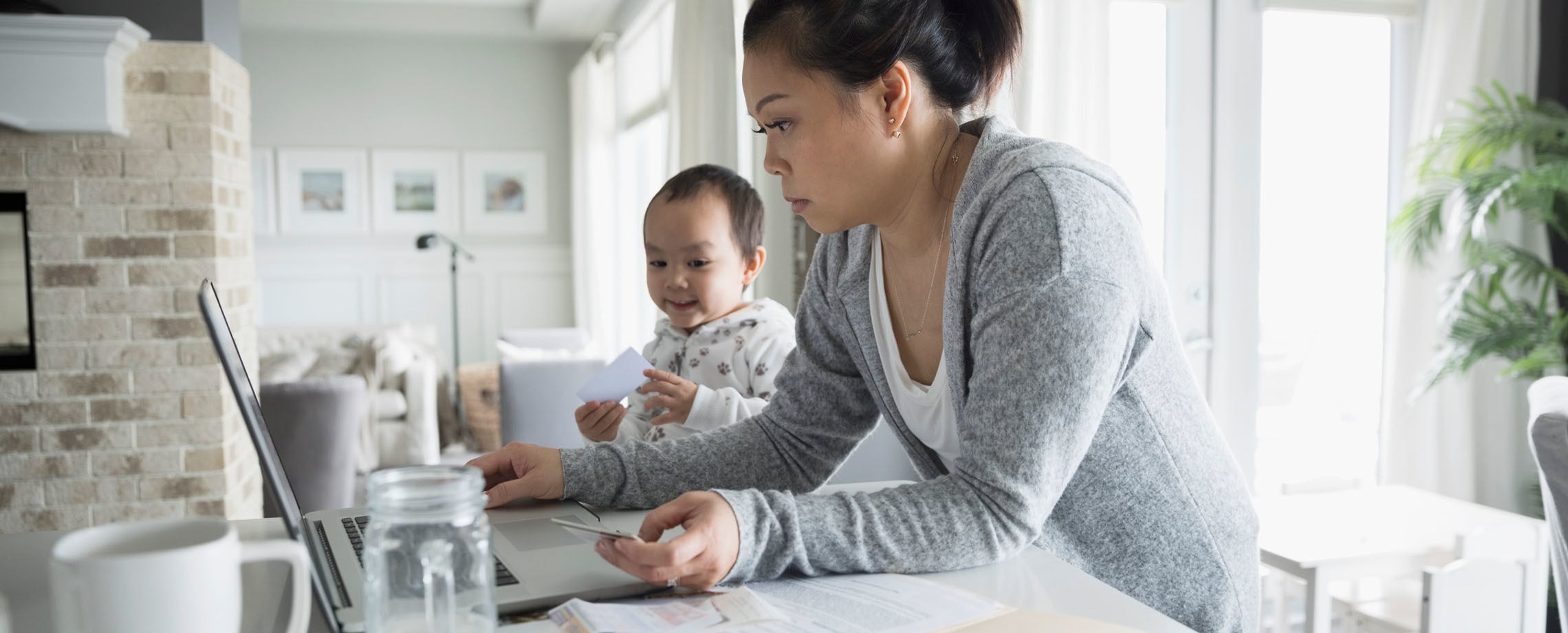 Asian-American woman holding plastic card and looking at laptop with her baby looking on