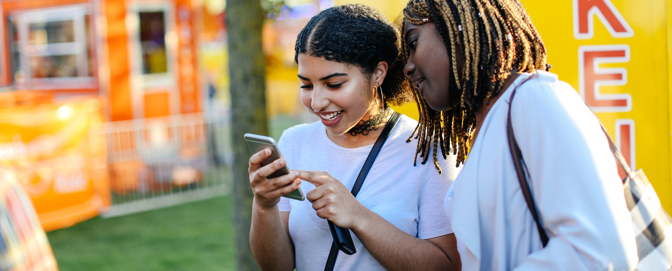 Two young African-American women looking at same cell phone while at outdoor festival