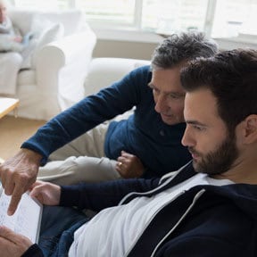 Retirement aged Caucasian man with grown son sitting on couch reviewing something on a tablet