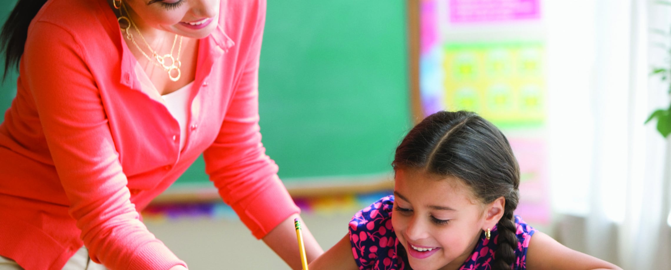 Close up of girl writing something in school as her teacher looks on, both smiling