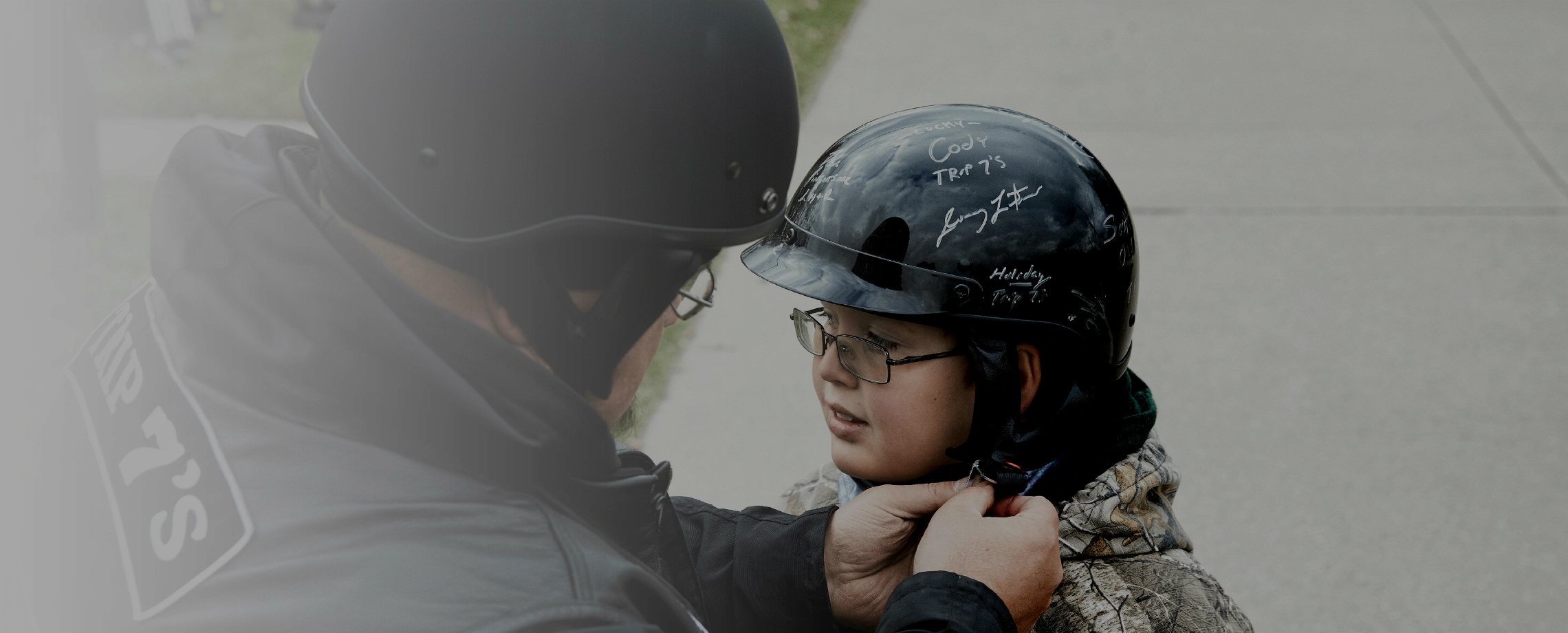 Caucasian man wearing leather jacket and motorcycle helmet helping youngster with his helmet