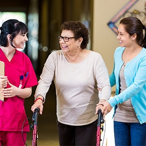 An older Hispanic woman using a walker taking to a nurse while being helped by her daughter