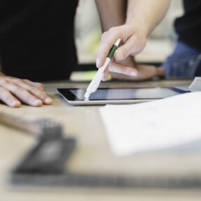 Hands using the back end of a pen to point out something on a tablet