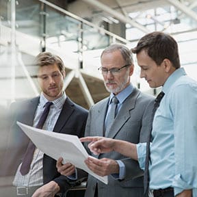 Three businessmen in suits and ties discussing a large sheet of paper in a factory