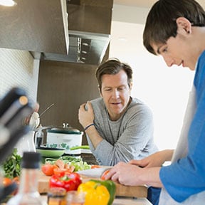 Man talking to his older teenaged son as the son cooks on the stove