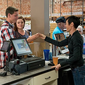 Young Asian man behind cash register smiling while handing something to a young Caucasian couple