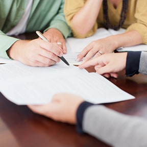 Closeup of a husband and wife's hands signing a document with other hands pointing where to sign