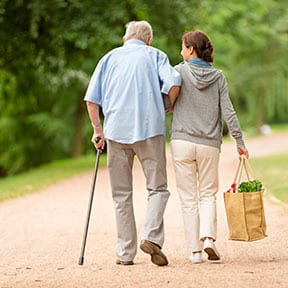 Older man with cane leaning on caregiver carrying a bag of groceries
