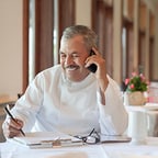 Man in chef's whites with reference papers using his mobile phone in empty dining room
