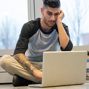 Young man with trendy haircut and tattooed arm sitting on floor and working on laptop