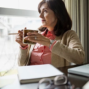Middle aged woman sitting by window holding pencil and coffee cup