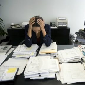 Businesswoman at a table covered with stacks of paper, holding her head in her hands