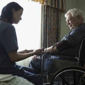 Nurse taking blood pressure of older grey haired man sitting in wheelchair