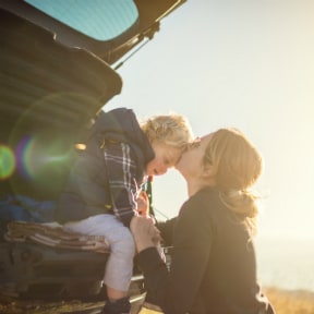 Woman kissing her toddler son's forehead as he sits on tailgate of vehicle