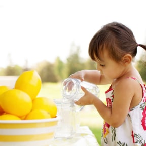 Very young girl pouring water from one mason jar to another with bowl of lemons on the table