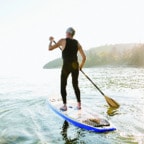 Caucasion woman with short grey hair wearing a black wet suit paddle boarding