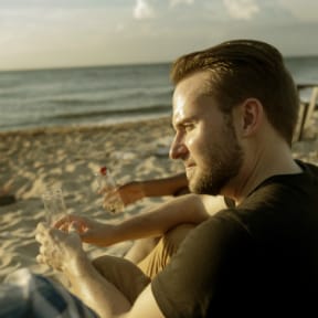 Bearded man sitting on the beach watching the ocean with an empty glass in  hand