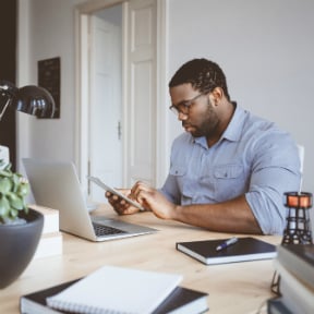 African-American thirty something male in home office looking at tablet with laptop also in front of him