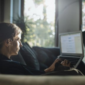 Young Caucasian man with long hair, sitting on couch looking at laptop