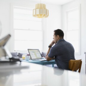 Man at kitchen table, looking at laptop and biting thumbnail