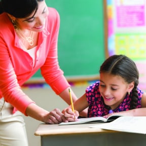 Girl writing something in school as her teacher looks on, both smiling